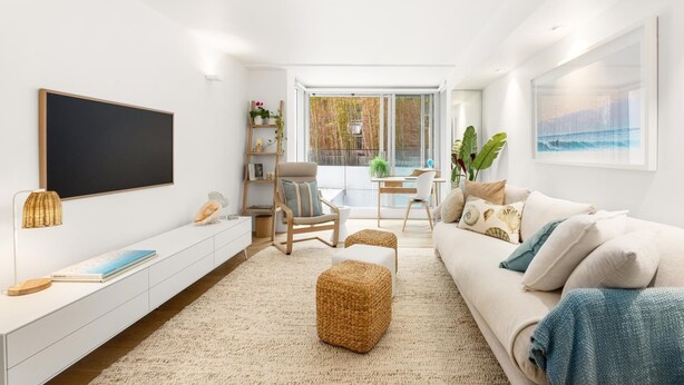 Una sala de estar moderna con un televisor de pantalla plana, un sofá blanco, una zona de comedor con mesa y sillas, y un ventanal con vistas a árboles.