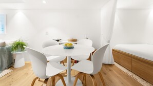 Una habitación moderna y minimalista con una mesa redonda blanca, cuatro sillas blancas, piso de madera, una cama blanca y una planta pequeña.
