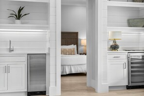 A modern hotel room with a kitchenette, a bed with white bedding, a nightstand with a lamp, and a potted plant on the shelf. - 30A Beach House - Dream by the Sea  (Santa Rosa Beach)