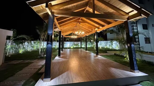 Un espace extérieur abrité, doté d’un plancher en bois et de colonnes, entouré de verdure et de bâtiments.