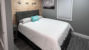 A modern bedroom with a bed, a nightstand, a wooden accent wall, a painting, and a window with blinds. - Charming 2-bedroom bungalow in Smithfield with AC, WiFi, washer and dryer (Smithfield)