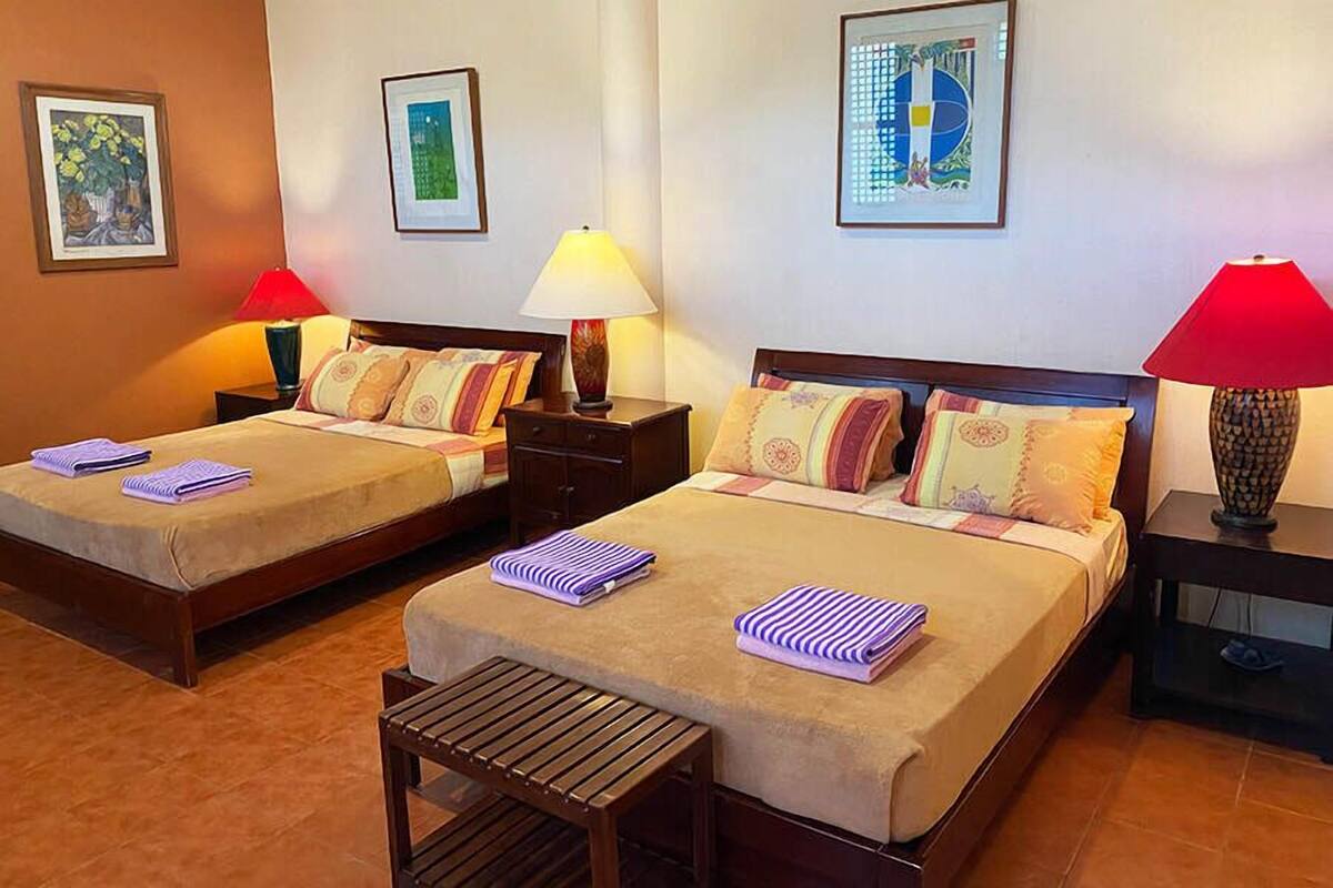 A hotel room with two beds, each with a wooden headboard, a small bedside table, and a lamp. There are framed artworks on the walls and a bench in the center of the room.