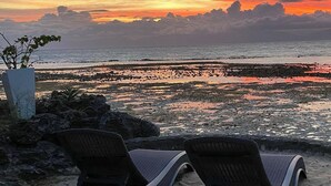 Two lounge chairs on a rocky beach with a view of the ocean and a sunset.