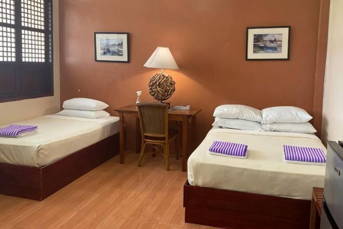 A hotel room with two single beds, a desk with a chair, a lamp, and two framed pictures on the walls.