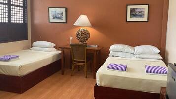A hotel room with two single beds, a desk with a chair, a lamp, and two framed pictures on the walls.