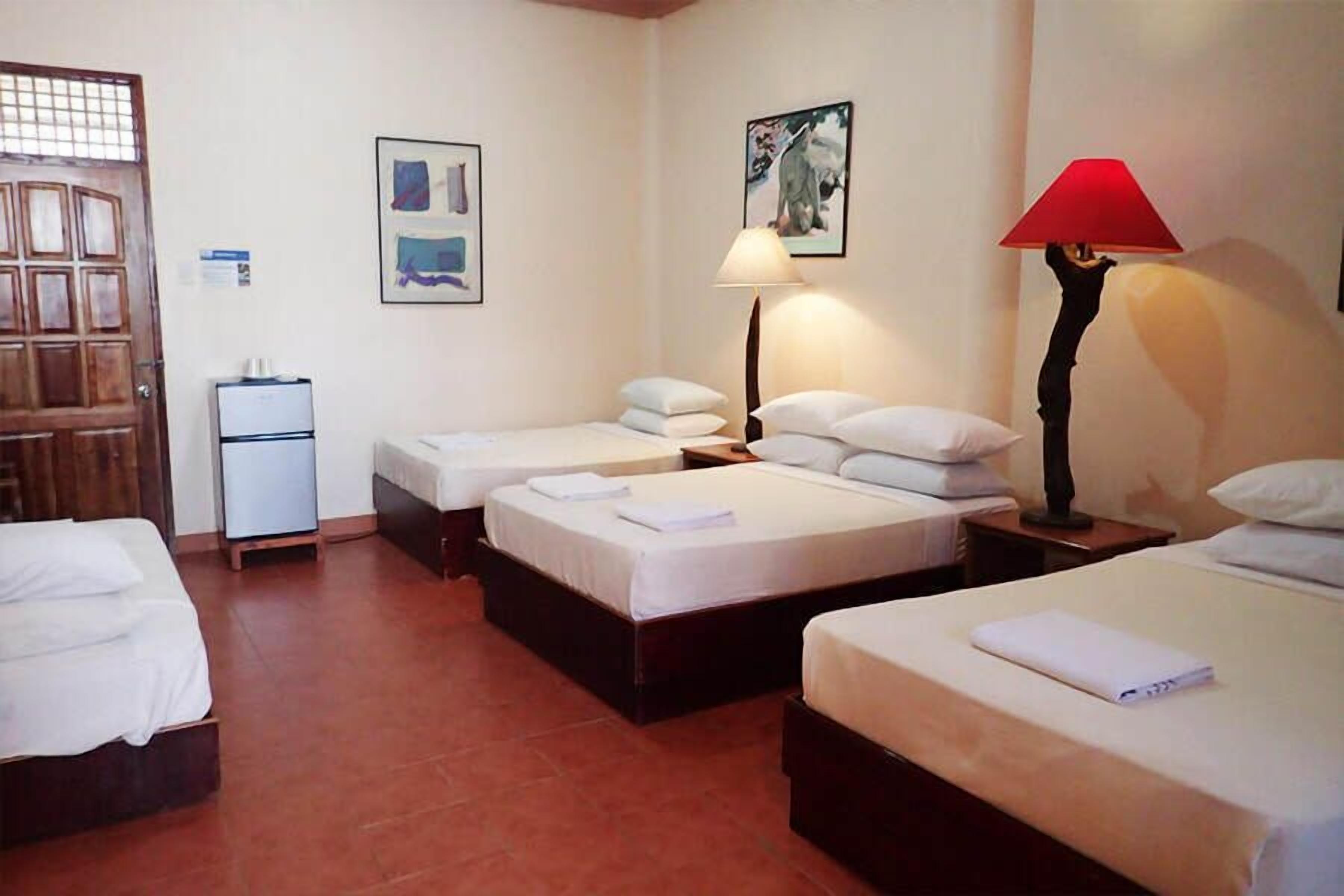 Une chambre d’hôtel avec trois lits simples, un petit bureau, un réfrigérateur et une lampe rouge.