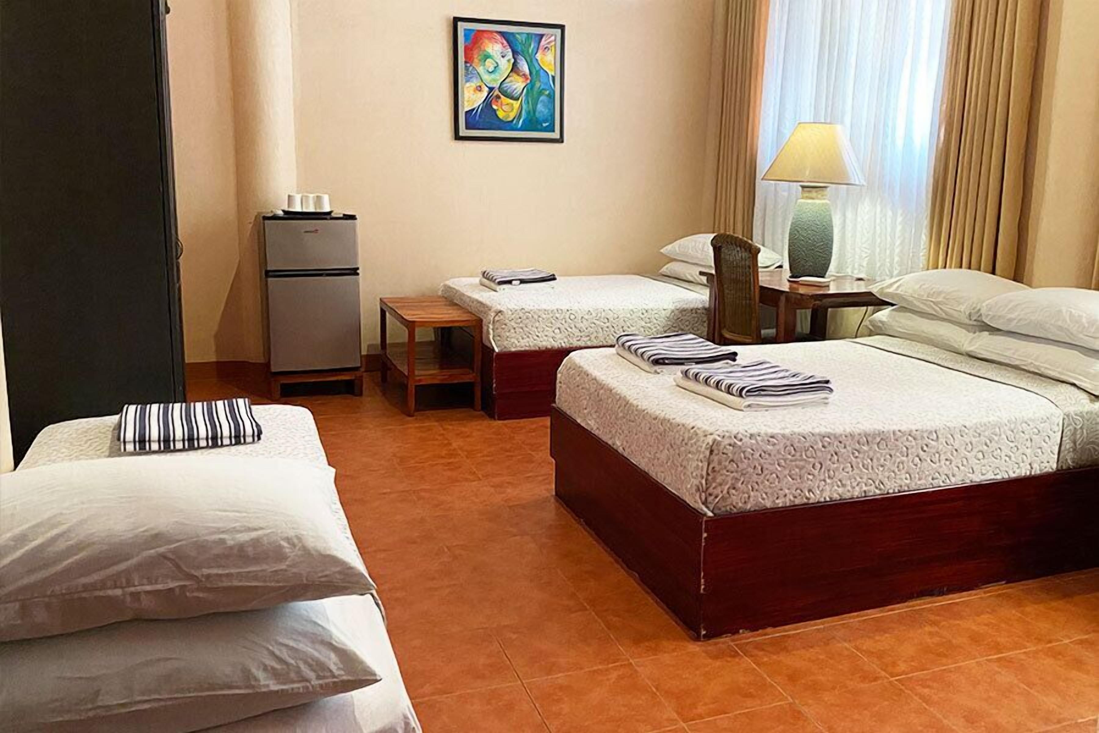 Une chambre d’hôtel avec deux lits, une petite table, une lampe, un réfrigérateur et un tableau au mur.