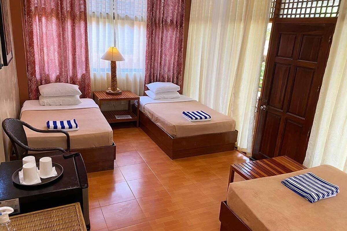 A hotel room with two beds, a small table with a lamp, a chair, and a small table with cups and a bottle.