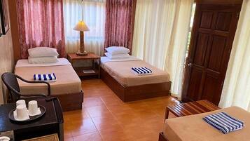 A hotel room with two beds, a small table with a lamp, a chair, and a small table with cups and a bottle.