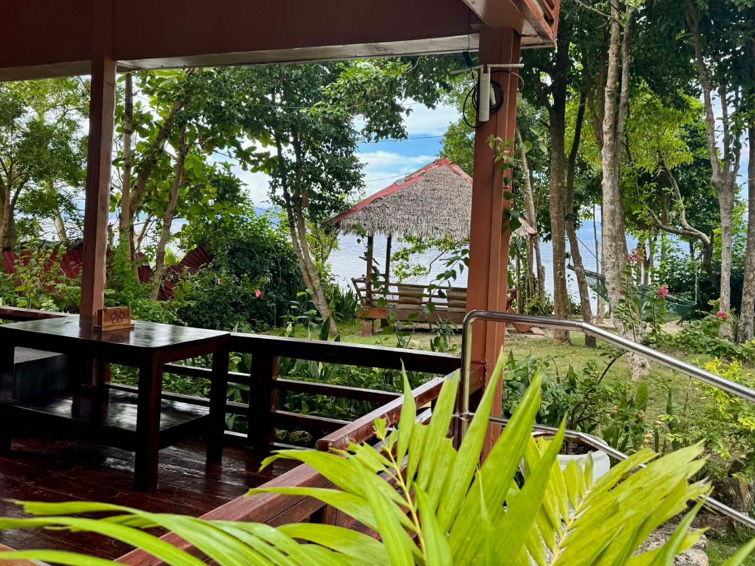 Une terrasse en bois avec une table et des chaises, entourée d’une végétation luxuriante et, au fond, une structure dotée d’un toit de chaume.