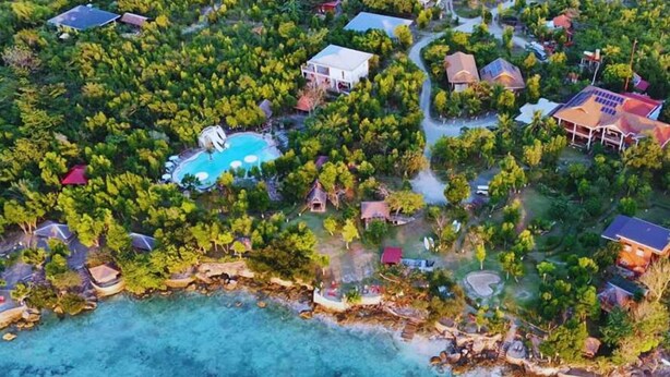 An aerial view of a coastal resort with multiple buildings, a swimming pool, and clear turquoise waters.