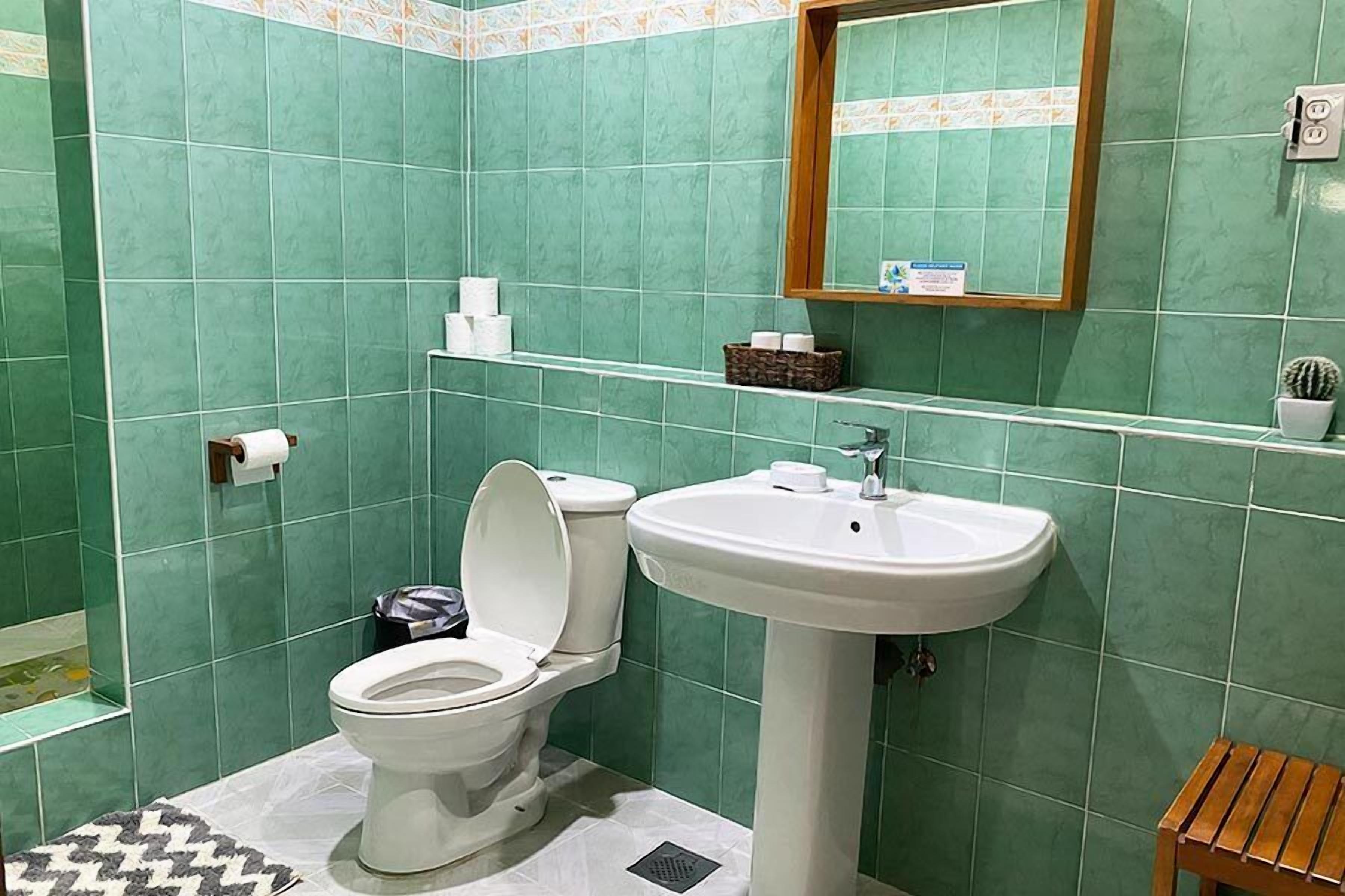 Un baño con azulejos verdes, un inodoro blanco, un lavamanos y un taburete de madera pequeño.