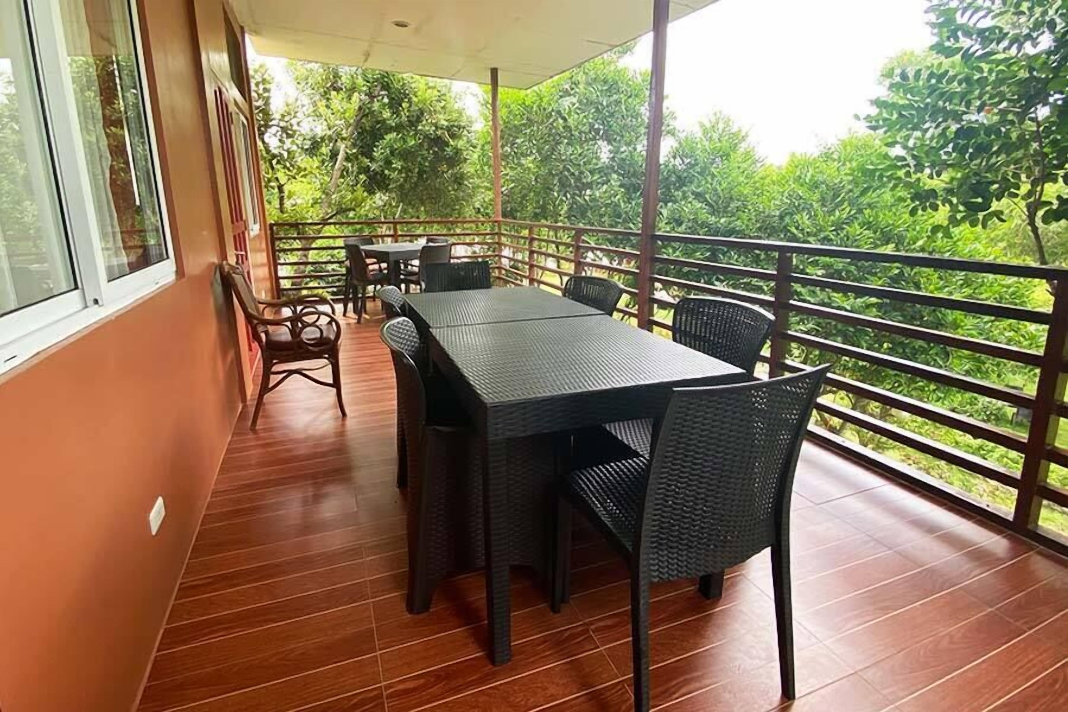 A covered outdoor seating area with a table and chairs, a wooden floor, and a view of greenery.