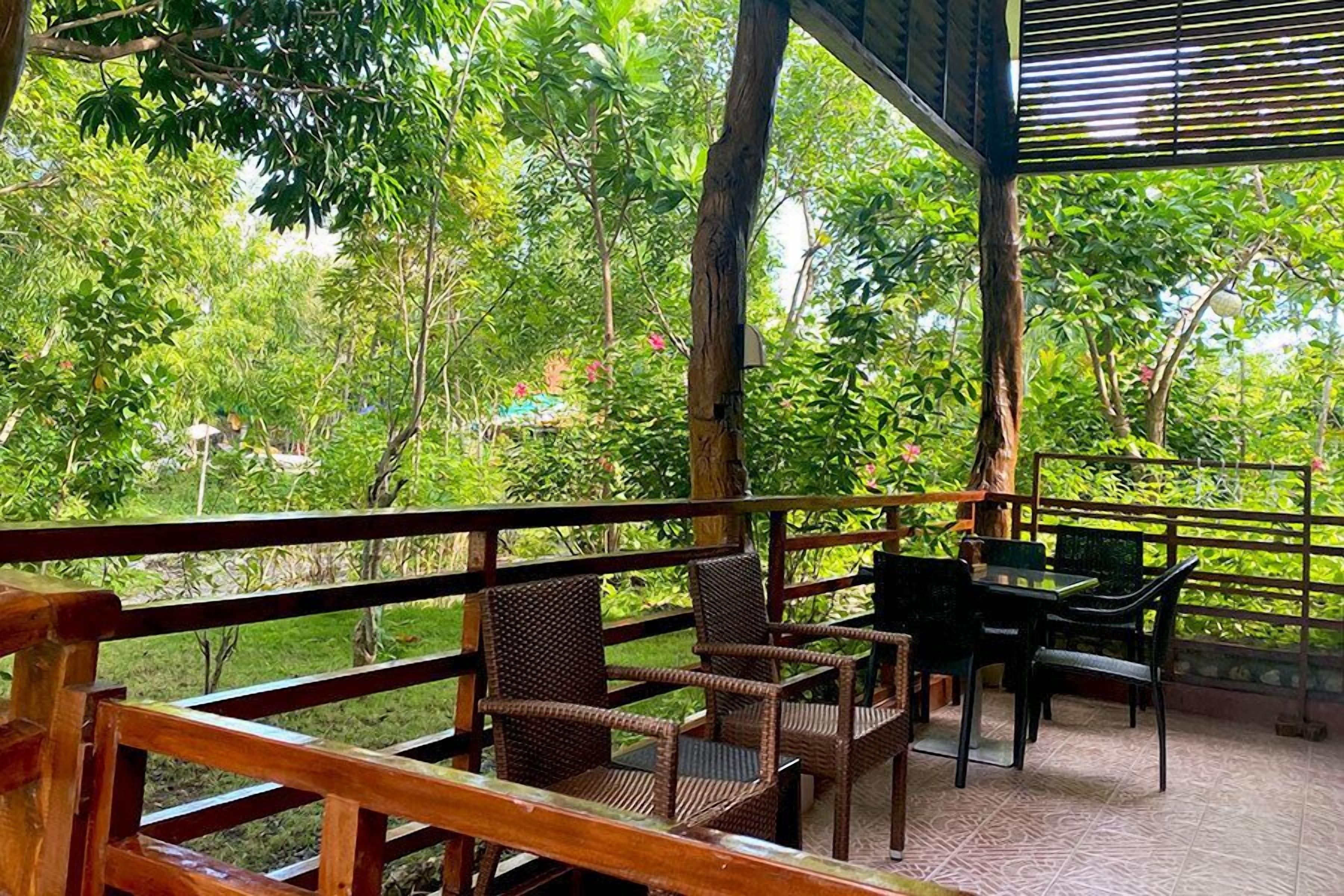 A covered outdoor seating area with wicker chairs and a table, surrounded by lush greenery and trees.