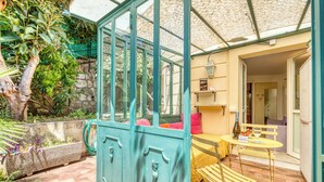 A conservatory with a glass door, a seating area with a yellow sofa and a small table, and a stone planter with plants.