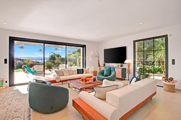 A modern living room with a large sliding glass door leading to a balcony, a flat-screen TV mounted on the wall, and a variety of seating options including a sofa, chairs, and a coffee table.