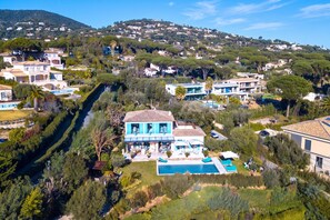 A residential area with a large house featuring a swimming pool, surrounded by lush greenery and a hilly backdrop.