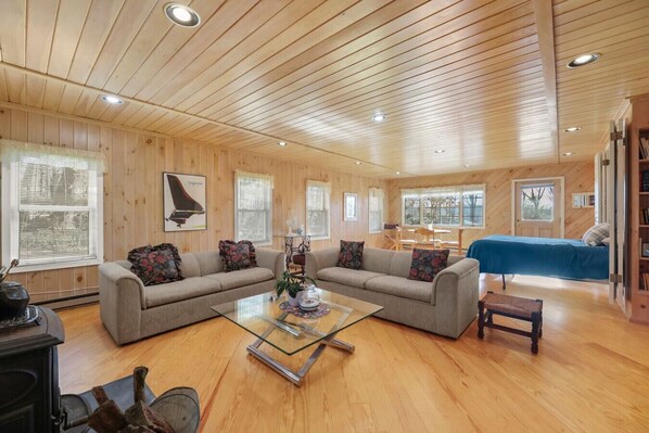 A cozy living room with a wooden ceiling, two sofas, a glass coffee table, and a blue bed in the background.
