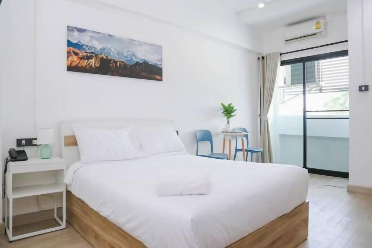 Een slaapkamer met een bed, een nachtkastje, een klein tafeltje met stoelen, een potplant en een raam met gordijnen.
