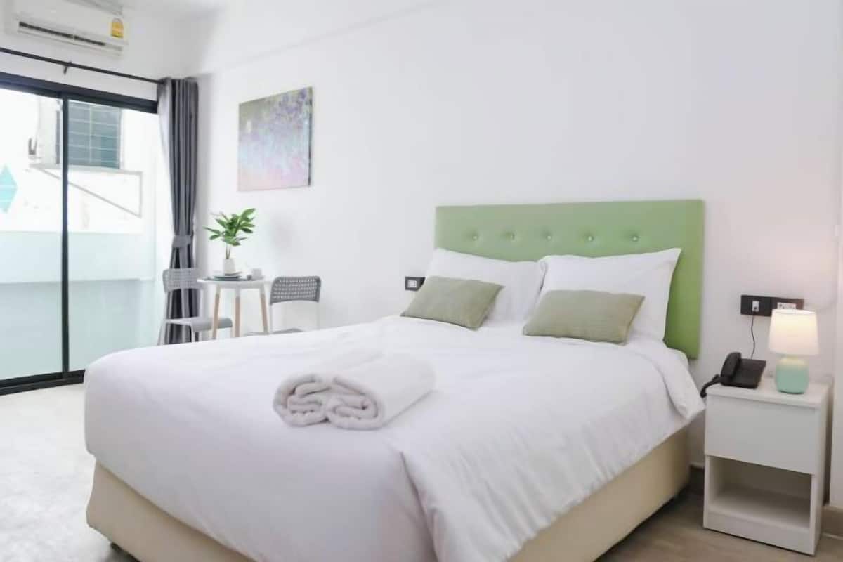 A hotel room with a bed, a bedside table with a lamp, a small table with a plant, and a window with a view.