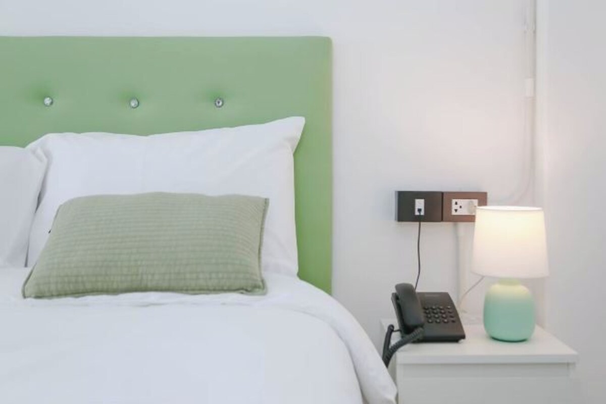 Un lit avec une tête de lit verte, une literie blanche, un oreiller vert, une lampe de chevet et un téléphone sur une table de chevet blanche.