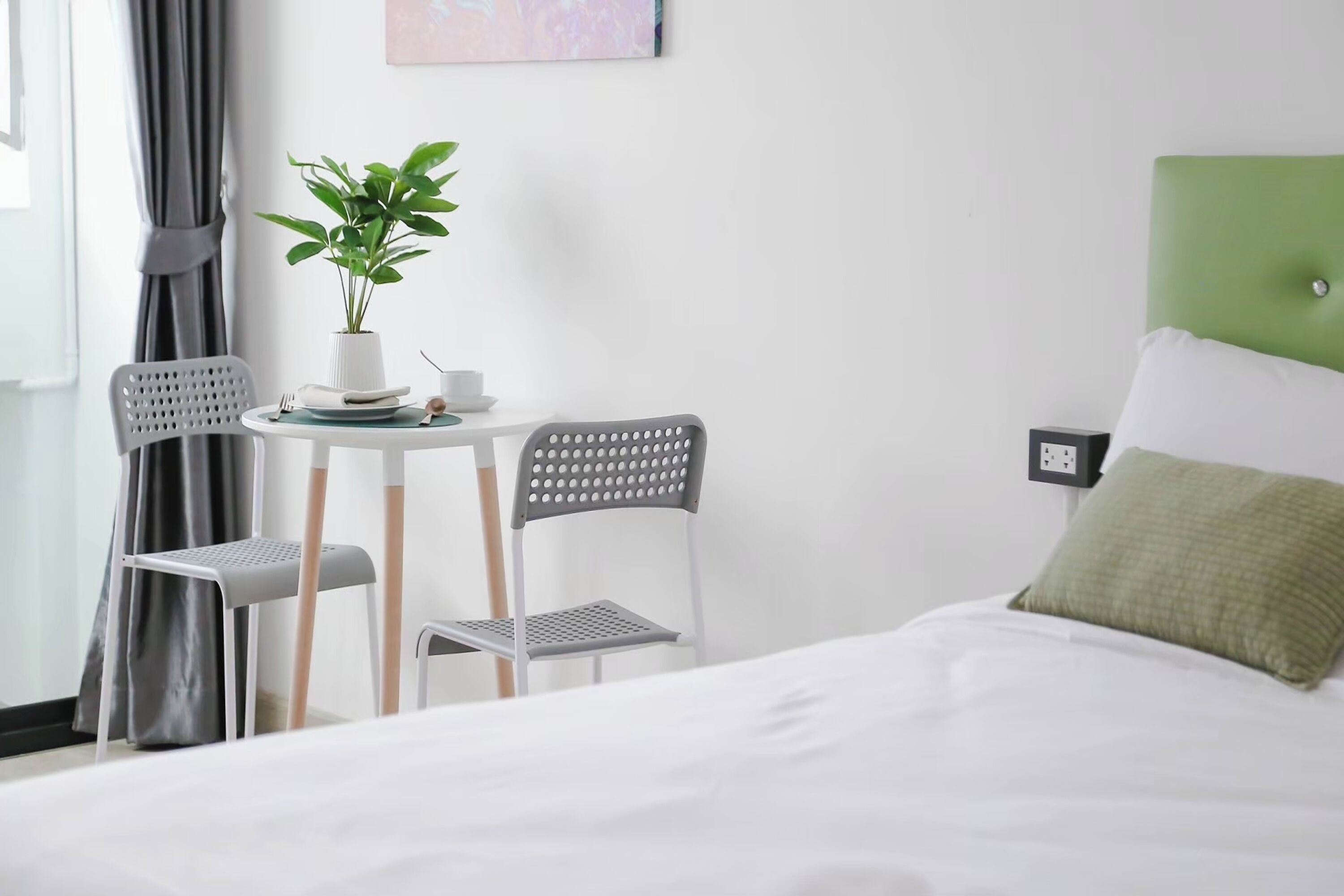 A neatly arranged hotel room with a bed, a small table with chairs, a potted plant, and a wall-mounted power outlet.