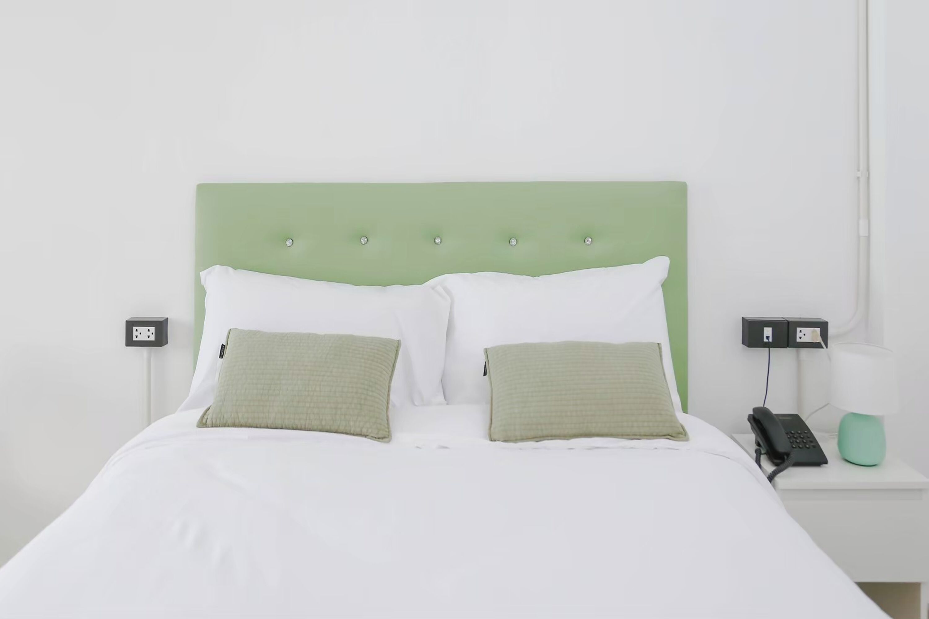 A neatly made bed with a green headboard, white bedding, and two green pillows. There is a bedside table with a lamp and a phone.