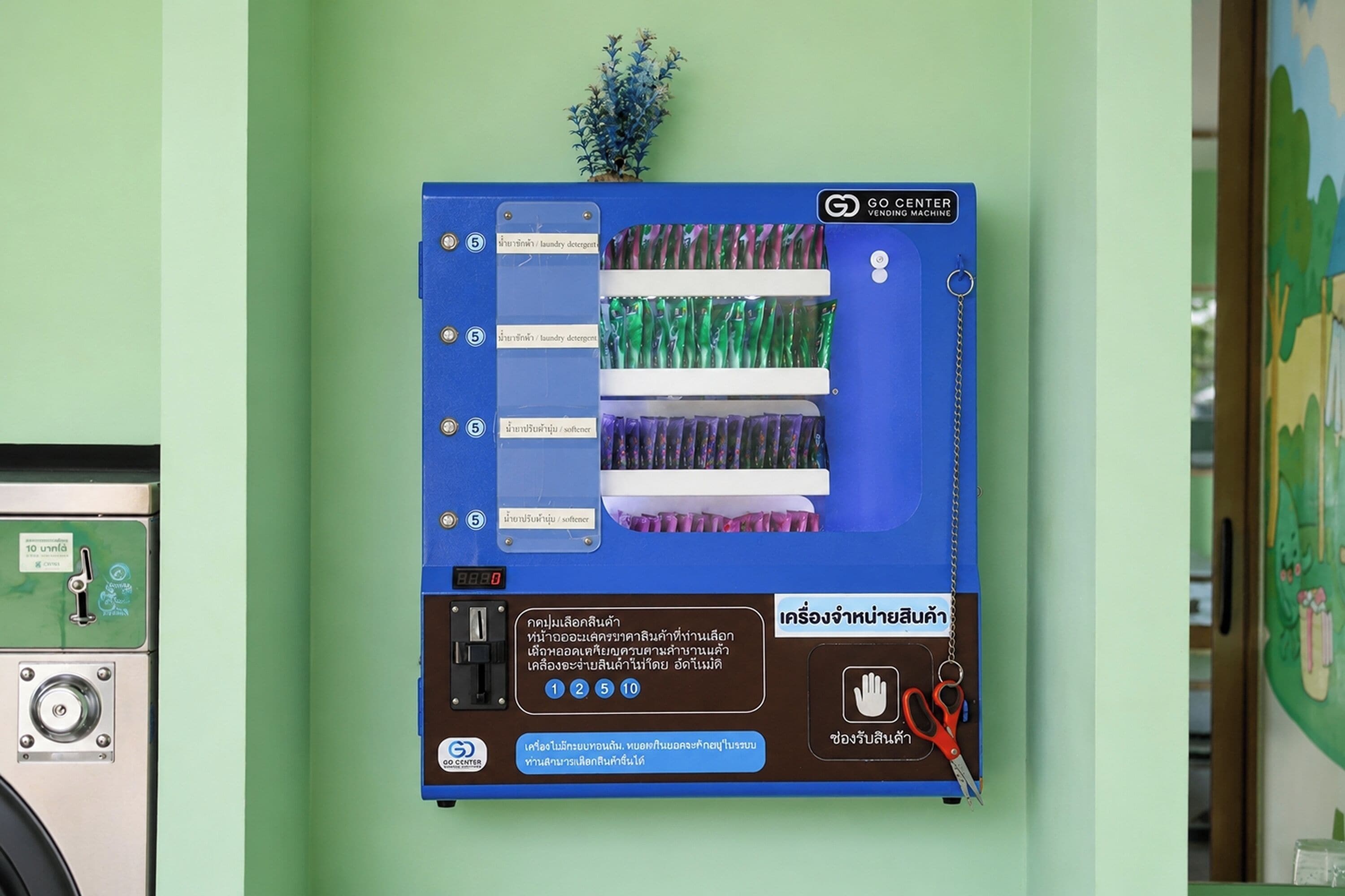 Una màquina expenedora amb diversos productes, una petita planta i un panell de control fixat a la paret.