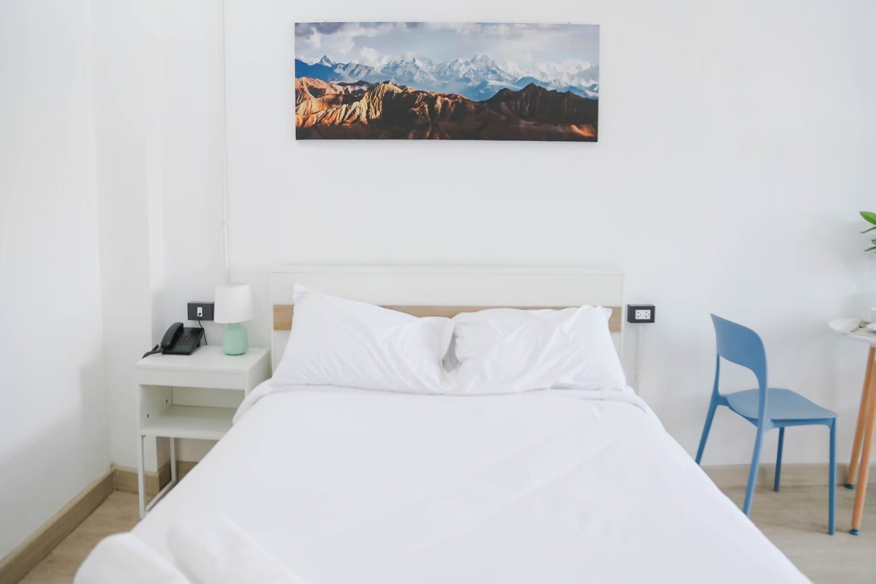 A hotel room with a bed, a desk with a phone, a chair, and a wall-mounted picture of mountains.