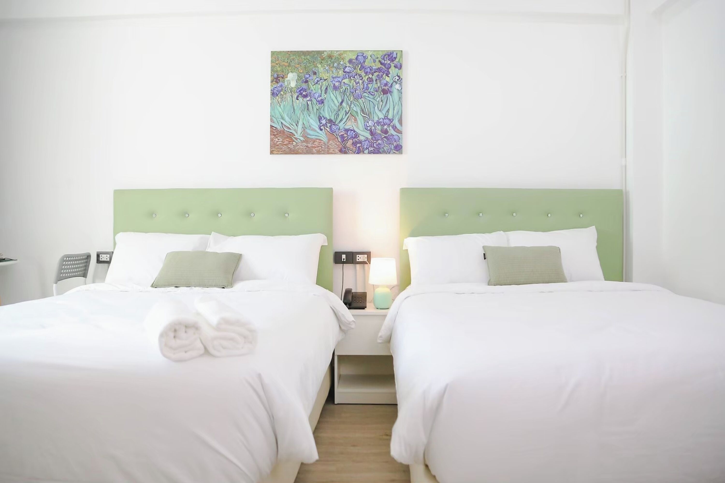 A hotel room with two beds, each with a green headboard, white bedding, and a green pillow. There is a painting above the beds, a bedside table with a lamp, and a phone on the table.