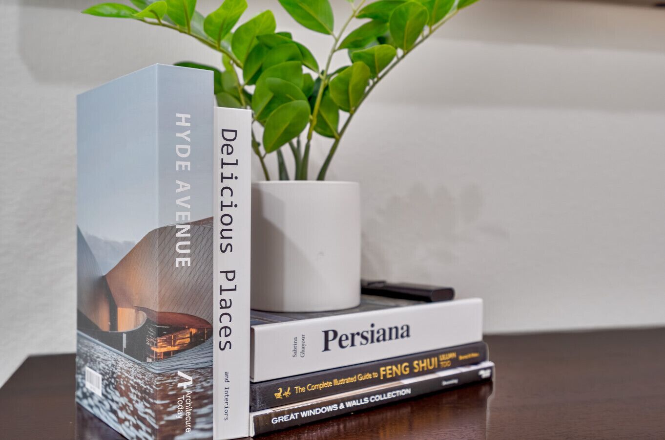 A stack of books titled 'Hyde Avenue', 'Persiana', and 'The Complete Illustrated Guide to Feng Shui' on a wooden surface with a potted plant in the background.