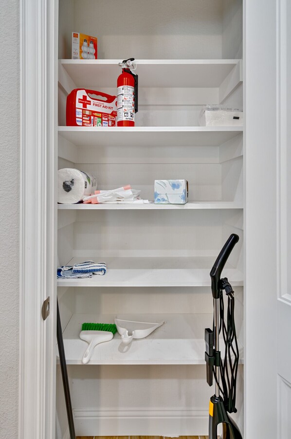 A white closet with shelves containing a fire extinguisher, toilet paper, cleaning supplies, and a broom.