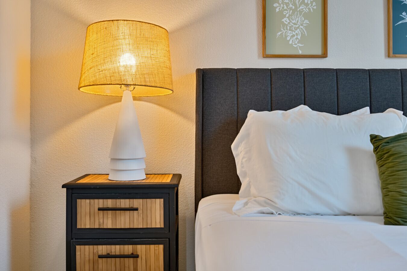 Un lit bien fait, avec du linge de lit blanc et un coussin vert, une table de chevet avec une lampe, et deux œuvres d’art florales encadrées accrochées au mur.