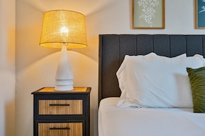 Ein ordentlich bezogenes Bett mit weißen Leinen und einem grünen Kopfkissen, ein Nachttisch mit einer Lampe und zwei gerahmte Blumenbilder an der Wand.