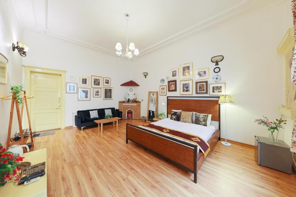 A bedroom with a wooden bed, a black sofa, a wooden dresser, and a small table. There are framed pictures on the wall and a chandelier above the bed.