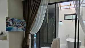A modern bathroom with a large bathtub, a shower area, and a sink. There is a television mounted on the wall and a window with curtains. - Baan First Pool Villa (Koh Samui)