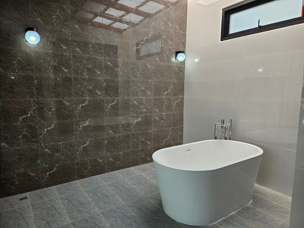 A modern bathroom with a freestanding bathtub, marble walls, and a small window.