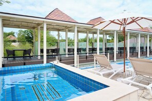 Een overdekt buitenzwembad met ligbedden en parasols, omgeven door een paviljoen met een betegeld dak.