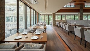Un restaurante con largas mesas de madera y sillas tapizadas de color gris, amplias ventanas que ofrecen vista a árboles, y un pilar de piedra en el centro.