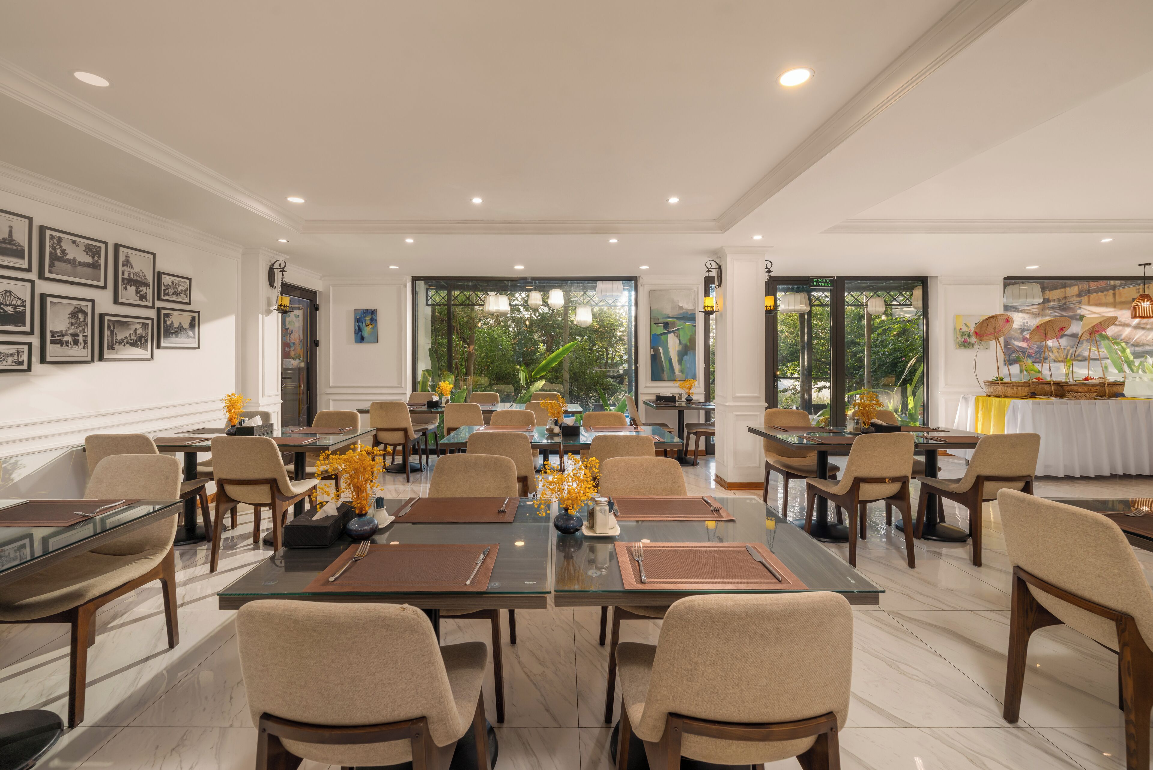 A spacious dining area with multiple tables and chairs, framed pictures on the walls, and large windows offering a view of greenery outside.