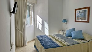 A bedroom with a bed, a nightstand, a lamp, a window with curtains, and a wall-mounted picture.