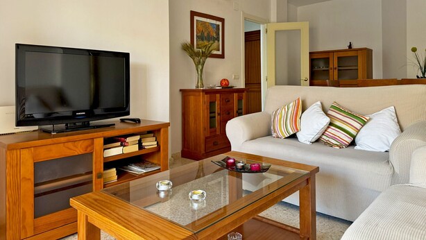 A living room with a flat-screen TV on a wooden stand, a glass coffee table, a sofa with striped pillows, a wooden cabinet, and an air conditioning unit on the wall.