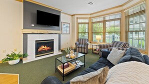 A living room with a fireplace, a television mounted on the wall, a sofa, a striped armchair, and large windows with blinds. - Breezy Beach | Villa 75 Steps from Lake Michigan (Sheboygan)