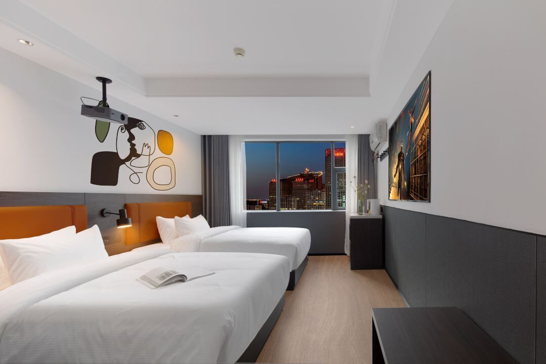 A modern hotel room with two beds, a desk, a large window with a city view, and a projector on the ceiling.
