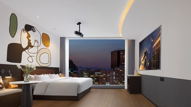 Een moderne hotelkamer met een groot bed, uitzicht over de stad, een beamer en een schilderij aan de muur.
