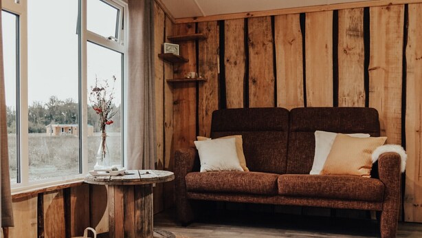 Ein rustikaler Raum mit einem braunen Sofa, einem runden Holztisch mit einer Vase und einem Fenster mit Blick auf ein Feld.