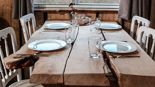 Una mesa de madera rústica para cuatro personas, con sillas blancas, una ventana con cortinas y vistas a árboles y un edificio.
