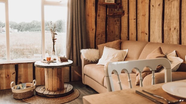 Une pièce chaleureuse au décor intérieur en bois, avec une table ronde en bois agrémentée d ’un vase et de livres, un canapé beige avec des coussins, et une vue sur un champ par la fenêtre.