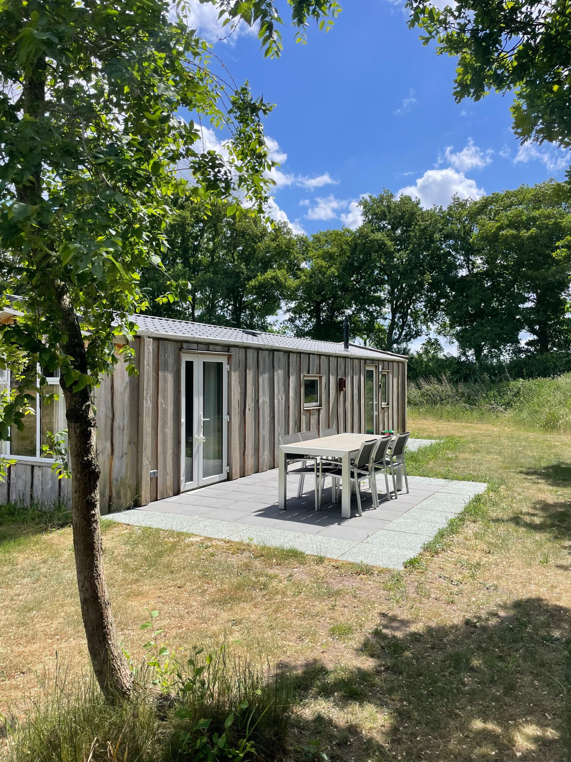 A wooden cabin with a patio table and chairs, surrounded by trees and a grassy area.