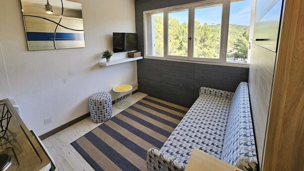 Un espacio habitable pequeño con un sofá, un televisor en un estante, un ventilador de techo y una ventana con vista a árboles.
